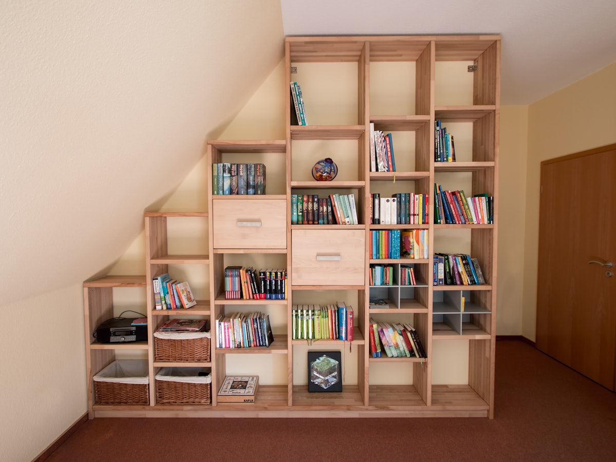 Bibliothèque en bois clair montée en gradins sous un rampant de comble, garnie de livres, de paniers en osier et de deux tiroirs.