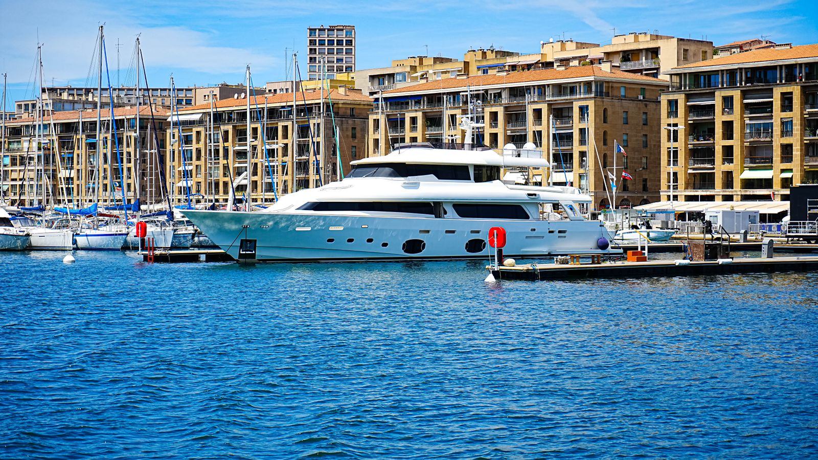 Un grand bateau blanc amarré au quai d’un port de plaisance, devant des immeubles en pierre claire aux toits de tuile.
